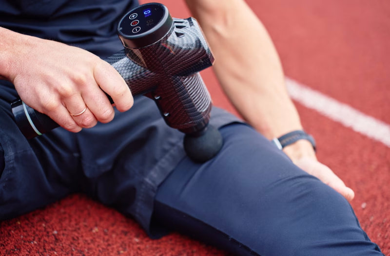 Person using a handheld massage gun on leg muscles after workout on track.